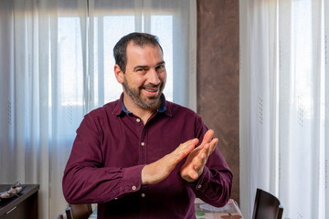 Young handsome man with a beard wearing a purple shirt clapping cheerfully, smiling and proud. Hands together