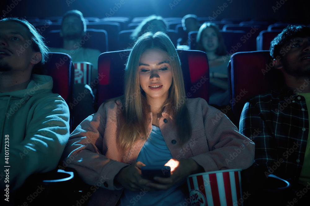 Beautiful young woman smiling, using her phone for texting while watching a movie at the cinema - obrazy, fototapety, plakaty 
