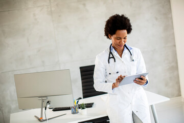 Female African American doctor wearing white coat with stethoscope standing by desk in office and looking at digital tablet