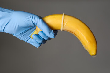 Wall Mural - Doctor holding banana in condom symbolizing male sexual organ on grey background, closeup. Potency problem