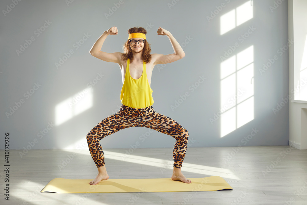 Thin athlete standing in funny pose and doing sports exercise during workout at home or at the gym. Happy guy in hilarious retro fitness wear smiling, looking at camera and showing weak arm muscles - obrazy, fototapety, plakaty 