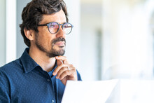 Business Man Wearing Glasses  Holding Paper Document Contract And Looking At The Window. Successful Male Portrait Thinking And Reading Contract At The Office