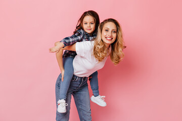 Wall Mural - Mom in white T-shirt and jeans plays with her daughter, holding her on back. Shot of adult and little girls having fun on pink background