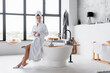 Woman in towel and bathrobe holding cosmetic cream in modern bathroom