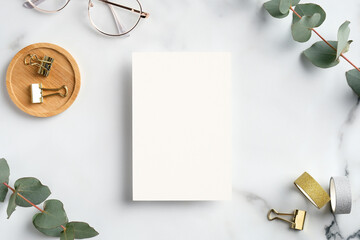 Wall Mural - Blank white greeting card with feminine accessories, office supplies and eucalyptus leaves on marble desk table. Flat lay, top view, copy space.