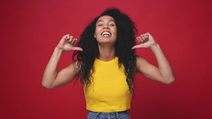 Wall Mural - Funny laughing african american young woman in yellow casual basic tank top posing isolated on red color background in studio. People sincere emotions lifestyle concept. Pointing thumbs on herself