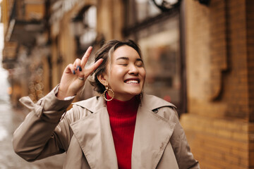 Positive girl in massive earrings laughing with closed eyes against background of building. Portrait of woman dressed in red sweater and beige jacket