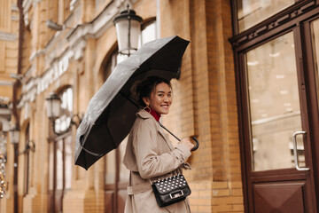 Beautiful woman in beige trench coat with cross-body bag with smile walks under umbrella in European city