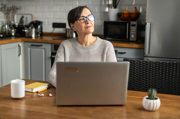 A thoughtful senior woman is sitting at the kitchen desk with a laptop, working remotely, takes a break and looks away searching inspiration and new ideas