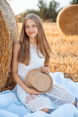 Canvas Print - Young smiling blond girl wearing white dress and straw hat, sitting on a white cloth in an agricultural field with straw bales..