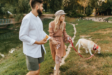 Hilarious husband and wife in stylish summer outfits walk along narrow path in park with their favorite dog