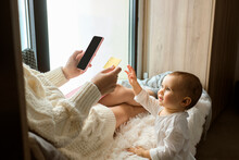Cute Baby Girl Reaching To Credit Card Being Held By Mother Sitting By Window At Home