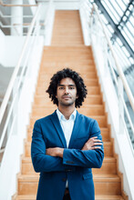Confident Handsome Young Male Entrepreneur Standing With Arms Crossed Against Staircase At Office