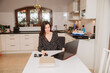 © Marianna - woman in kitchen. Beautiful woman is talking on the phone and working on the computer from home. Work during the pandemic. Woman in home office. The kitchen serves as an office