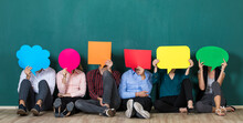 Group Of Six Business People Team Sittiing Together And Holding Colorful And Different Shapes Of Speech Bubbles Over Their Faces.
