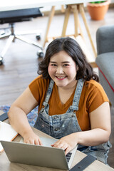 portrait of happy fat asian woman smiling and sitting on a floor typing on a computer laptop keyboard looking at camera from high angle view in a room. Working and study from home concept