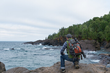 Male photographer wearing hiking backpack with camera tripods and gear strapped to it; looking out towards lake, rocky shore, and forest