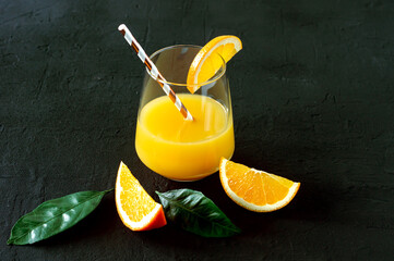 Wall Mural - A glass of juice and cut oranges with leaves on dark background