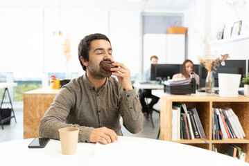 Hispanic man with mustache eating and enjoying a chocolate sweet bun. Unhealthy habits in the office concept.