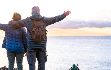 Rear view of two senior people enjoying the sea view and the freedom at sunset light.  Standing on the cliff looking at the horizon over water