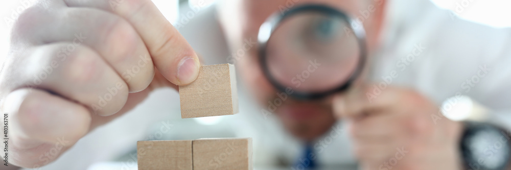 Close-up of man looking on wooden cubes stack through magnifying glass. Concentrated person building pyramid on working table. Business strategy and smart decision concept - obrazy, fototapety, plakaty 