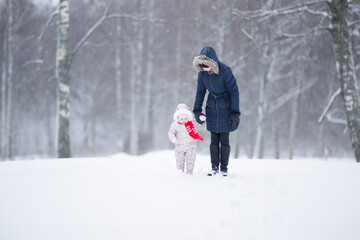 Wall Mural - Little daughter in overalls and young adult mother walking on white first snow at park. Spending time together in beautiful winter day. Enjoying peaceful stroll. Front view.