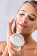 © LIGHTFIELD STUDIOS - woman with bare shoulders holding jar and applying face cream on white