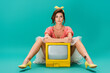 young pin up woman looking at camera while sitting near bright yellow vintage tv on turquoise