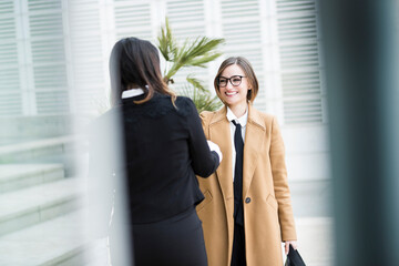 colleghe manager si incontrano in contesto business in esterni vicino ad un palazzo moderno fatto di specchi: Parlano entrambe al telefono mentre camminano