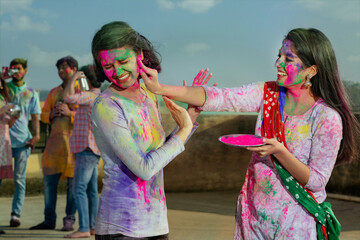 A YOUNG WOMAN PUTTING GULAL ON ANOTHER WOMAN WITH FRIENDS IN BACKGROUND	