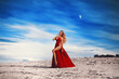 A gorgeous blonde girl in a sexy red dress with deep slits on the hips against the background of a white sandy beach and a sunset sky.