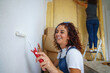 Decorating a new house and moving day concept. Happiness starting a new life with friends and family. Horizontal view of cheerful caucasian woman painting a white wall in her new apartment.