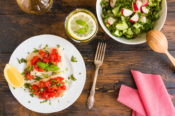 Halibut fish fillet baked with tomatoes on white plate. Salad with radish and cucumber in bowl. Wooden rustic table, top view