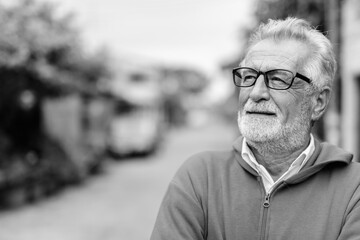 Wall Mural - Close up of happy handsome senior bearded man smiling while thinking and looking up with eyeglasses outdoors