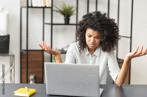 Young African American female office worker manager feeling stressed, tired, and overworked, does not know how to fix a problem, sitting at the desk in front of laptop, struggling and raising hands up