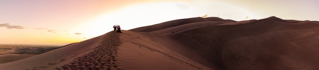 Panorama shot of group of people standing on top of sandy dune and going to sandskying down at sunset