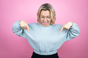 Wall Mural - Young caucasian woman wearing sweatshirt over pink background surprised, looking down and pointing down with fingers and raised arms