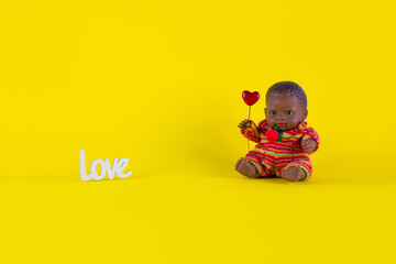 Happy Valentine's Day greeting card - black doll boy sits on a yellow background with a heart in his hands. Space for inscriptions