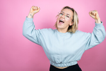 Wall Mural - Young caucasian woman wearing sweatshirt over pink background very happy and excited making winner gesture with raised arms, smiling and screaming for success.