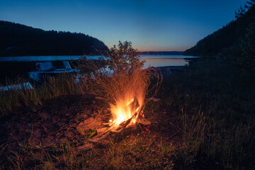 Wall Mural - A large night fire on the river bank in a forest clearing, flames, sparks, blue sky, light beyond the horizon, a boat near the shore.