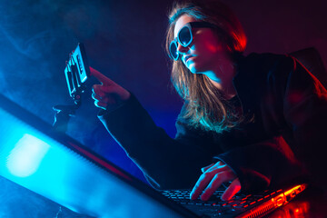 Poster - Geek woman next to a laptop. She uses a cell phone. Portrait of a geek woman in a dark room. Geek woman face is illuminated with neon light. Young girl works with a computer. She prints something