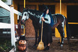 A girl dressed as a witch stands by a corral on a farm next to a horse