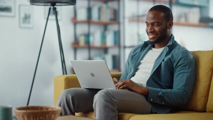 Wall Mural - Handsome Black African American Man Working on Laptop Computer while Sitting on a Sofa in Cozy Living Room. Freelancer Working From Home. Browsing Internet, Using Social Networks, Having Fun in Flat.