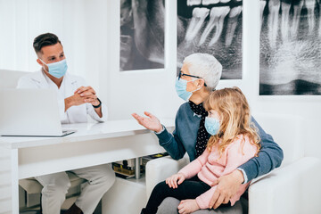 Wall Mural - Senior woman and her granddaughter sitting and talking with dentist at dentist's office. They are all wearing protective face masks due to coronavirus pandemic.