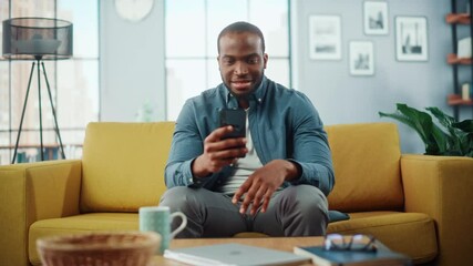 Aufkleber - Excited Black African American Man Using Smartphone while Resting on a Sofa in Living Room. Happy Man Smiling at Home and Chatting to Colleagues and Clients Over the Internet. Using Social Networks.