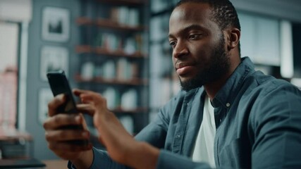 Aufkleber - Excited Black African American Man Using Smartphone while Sitting at Table in Living Room. Happy Man Smiling at Home and Chatting to Colleagues and Clients Over the Internet. Using Social Networks.