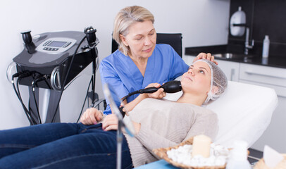 Mature female doctor cosmetologist performing machine facial procedure to young woman in cosmetic office