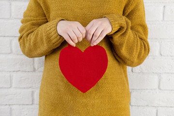 A young Caucasian woman holds a red heart in her hand against a white brick wall. Heart close-up. Valentine's Day. The concept of love, happiness, charity