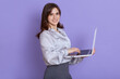 © sementsova321 - Portrait of adorable smiling woman with dark hair holding laptop isolated over lilac background, looks art camera, working via note book and wireless Internet, wearing elegant blouse and skirt.