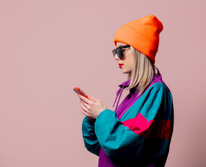 Wall Mural - Style girl in 80s sportsuit and sunglasses hold mobile phone on pink background